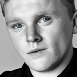 Black and white close-up portrait of a person with light-colored eyes, short hair, and a neutral expression, wearing a dark shirt.