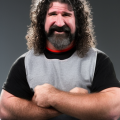 a person with long, curly hair and a beard stands with arms crossed, wearing a gray and black shirt against a gray background.