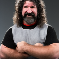 A person with long, curly hair and a beard stands with arms crossed, wearing a gray and black shirt against a gray background.