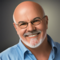 a bald man with glasses and a white beard smiles while wearing a light blue shirt against a grey background.